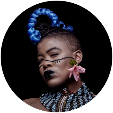 Headshot of Singer Thandiswa Mazwai looking toward the camera, donning a large blue braid, face markings, a flower earring and a blue and white beaded necklace against a black background.