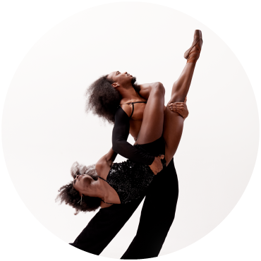 A dancer tilts their head up as they hold another dancer upside down dressed in black against a white background.