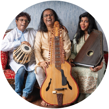 Subhasis, Debashish and Anandi Bhattacharya smile on a red couch and hold a drum, slide guitar, and swarmandal (small harp) against a blue background.