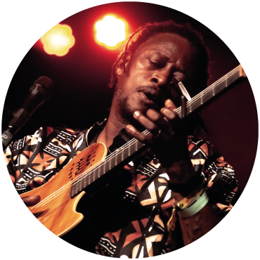 Habib Koité strums the guitar on stage while wearing a patterned shirt with orange lights in the background.