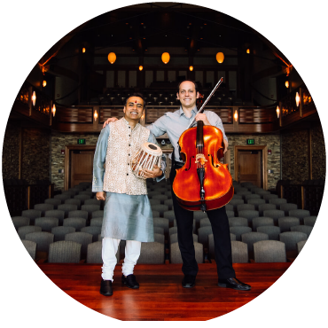 Sandeep Das holds a tabla and wears traditional Indian clothing next to Mike Block in black pants and a button-up who holds a cello. The duo are on front of the stage and the rest of the auditorium is visible.