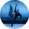 A male dancer stomps his left foot down and throws his arm in the air while on a stage lit by subtle blue lighting.