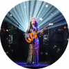 Marisa stands center stage wearing a sparkling dress and tiara. She holds a brown guitar. Spotlights shine from behind her up to the sky, creating a halo effect