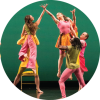 Two dancers dressed in pink and green cross hands on stage as they are held up by other dance members in front a green background.