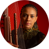 Headshot of Sona Jobarteh posing behind a kora in front of a red background.