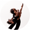 A dancer tilts their head up as they hold another dancer upside down dressed in black against a white background.