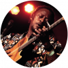 Habib Koité strums the guitar on stage while wearing a patterned shirt with orange lights in the background.