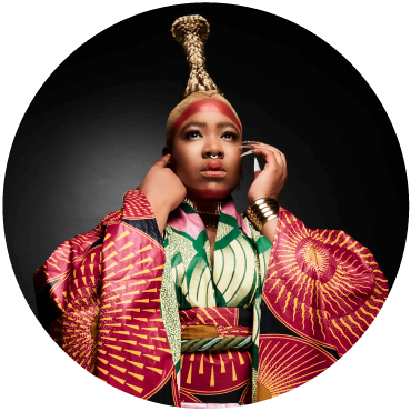  Singer Thandiswa Mazwai stares off into the distance wearing a large blonde braid, red face paint, and a kimono with vibrant african print.