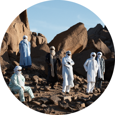 6 members of the band wearing full scarfs stand in front of large rocks in the desert