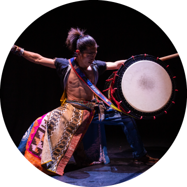 A yamato performer dressed in a patterned garb squats as he is about strike the drum sitting on his lap.